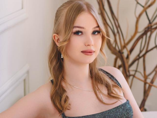 NicoleBrian with blonde pigtails, blue eyes, and subtle makeup, wearing a glittery green dress, small earrings, and a delicate necklace, has a cute, confident expression.