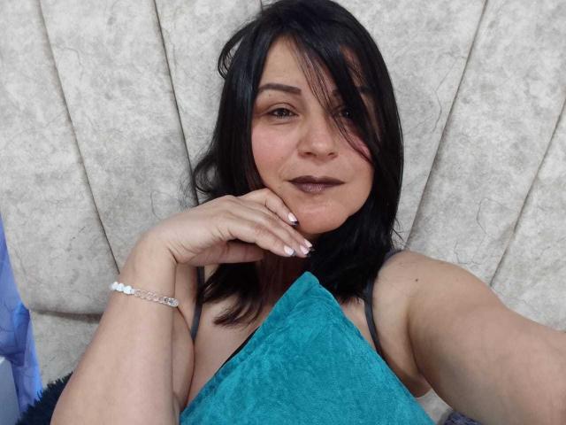 SexyAmature with black hair, medium tits, and dark lipstick, resting head on hand, wearing a teal pillow, white bracelet, and black top, with a relaxed, confident expression.