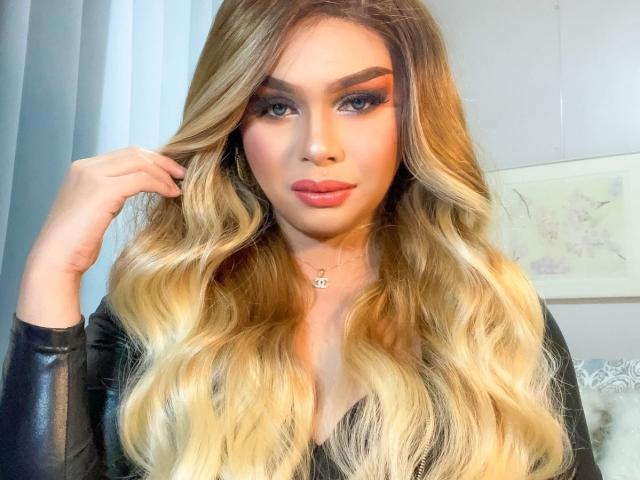 GoddessRoxy69 with long, wavy blonde hair, full eyebrows, heavy eyeliner, and pink lipstick, wearing a black leather top, holding her hair. She's got a cute, sexy face.