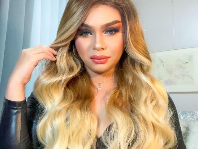 GoddessRoxy69 has blonde wavy hair, full red lips, and is wearing a black leather top. Her hand is in her hair. She has thick eyebrows and long eyelashes.