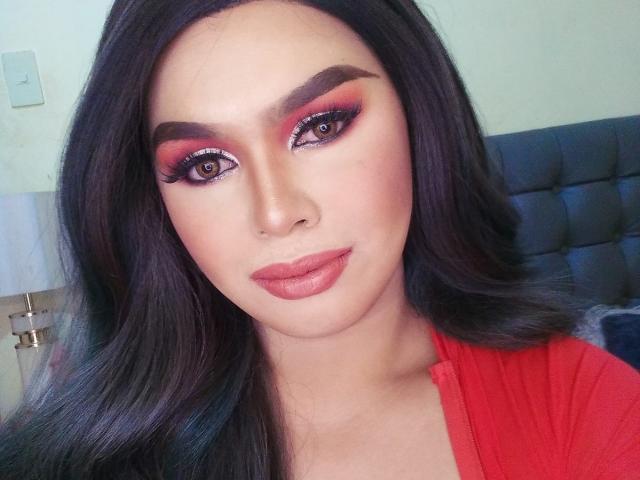GoddessRoxy69 has long black hair, red eyeliner, and pinkish-red lipstick. She's wearing a red shirt, looking at the camera with a slight smile. Her eyebrows are thick and dark.
