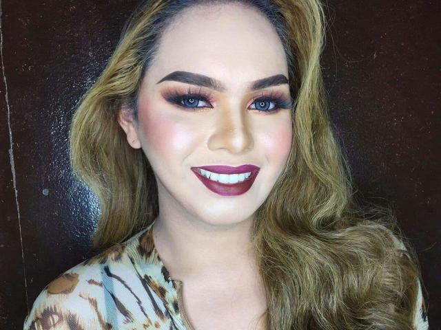 GoddessRoxy69 has blonde hair, blue eyes, thick eyebrows, dark red lipstick, and is wearing a tiger-print shirt. Her smile shows white teeth, and she has a sexy look.