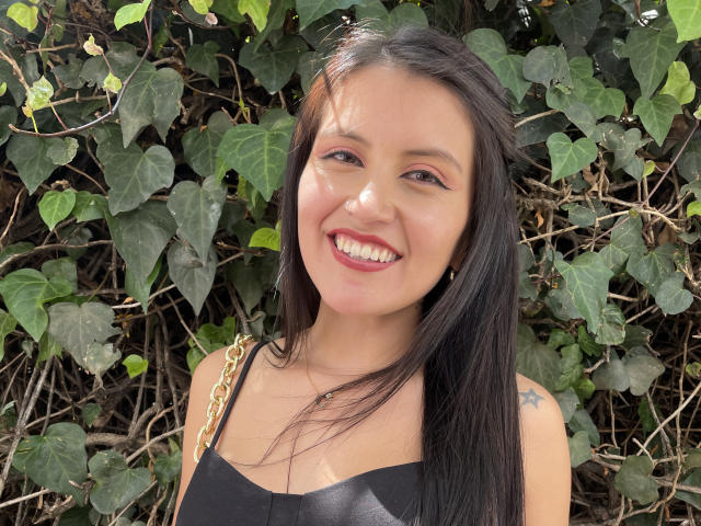 AmelieMuller with long black hair, smiling with red lipstick, wearing a black top with gold chain straps, and a small star tattoo on her right shoulder.