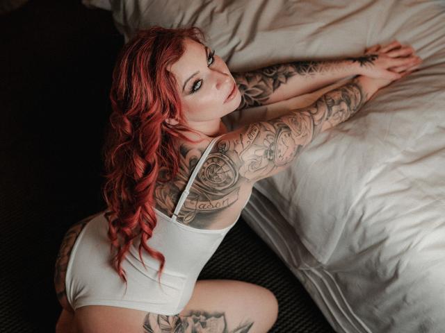 Chrissi69-hot with red hair, tattoos, white one-piece, kneeling on bed, looking back, hands on bed, showing ass and tattoos, in soft light.