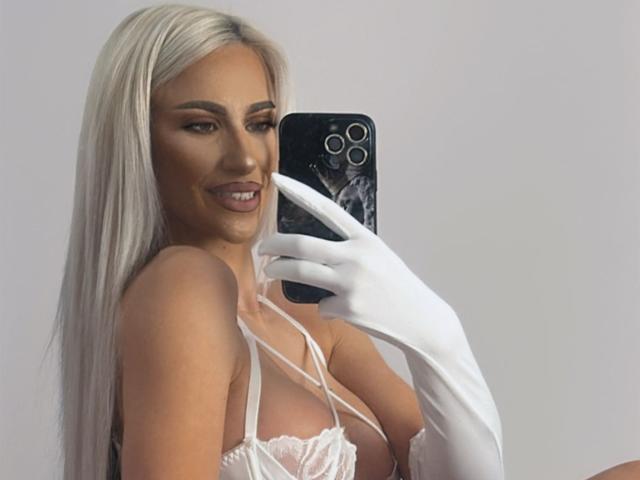 KarlaBart with long platinum blonde hair, wearing white gloves, taking a selfie with her phone, showing her bare tits in a white lace top, smiling.