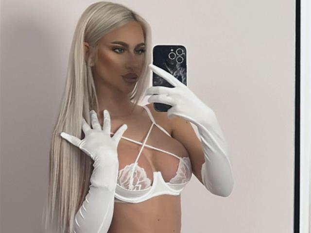 KarlaBart mit langen blonden Haaren, die ein weißes Spitzen-BH, ein trägerloses Top und weiße Ellbogenhandschuhe trägt, macht ein Selfie vor einem Spiegel mit ihrem Handy und präsentiert dabei ihre Brüste..