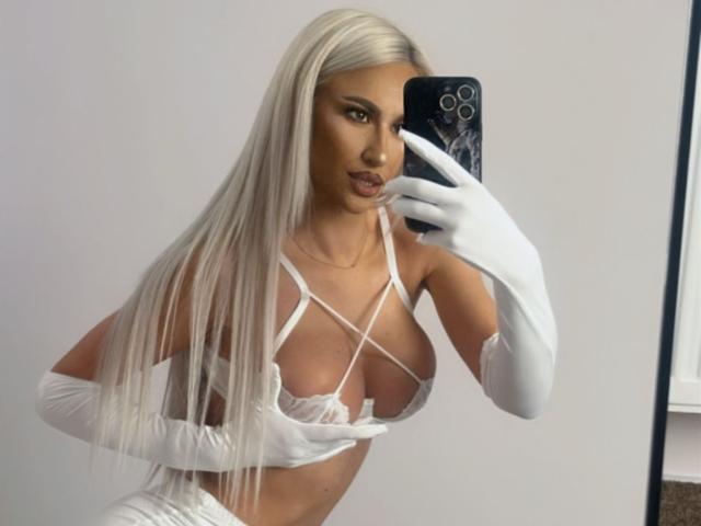 KarlaBart with long platinum blonde hair, wearing white strappy lingerie, long white gloves, and holding a phone, showing her big tits in a mirror selfie.