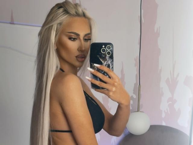 KarlaBart, con lunghi capelli biondi, indossa un top da bikini nero, tiene in mano un telefono con una custodia in marmo, e si mostra in un selfie allo specchio, mettendo in evidenza il seno e i glutei..