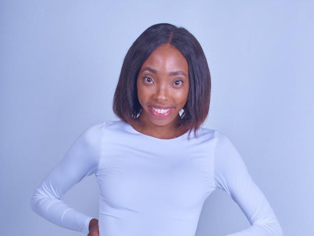 SlimPear69 has dark brown skin, straight black hair, and is wearing a white long-sleeve shirt, smiling with hands on hips. Her tits are small, and she's cute.