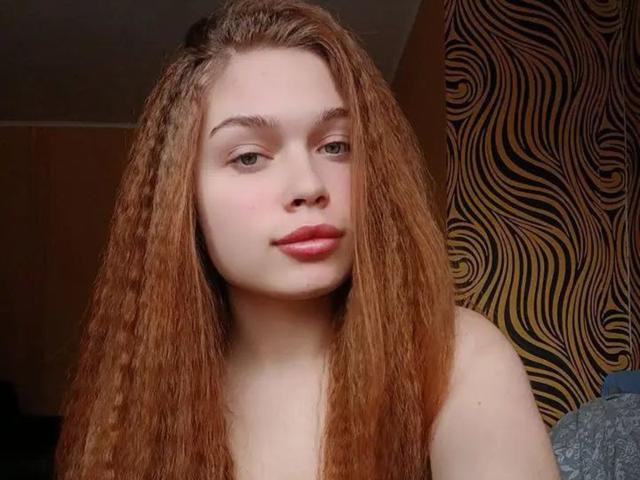 LaraPika with long, wavy red hair, pale skin, and pink lips, looking slightly to the side with a subtle smile, in a casual, topless selfie.