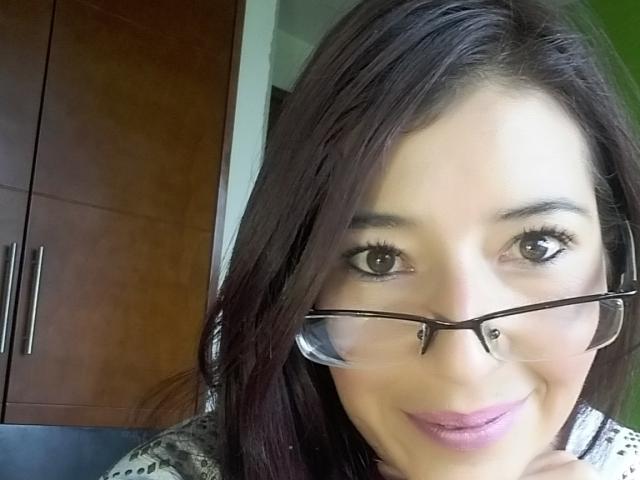 AntonnellaS with dark brown hair, glasses, and a slight smile, showing her face and pink lipstick. Her brown eyes and subtle makeup are visible. She's wearing a gray top.