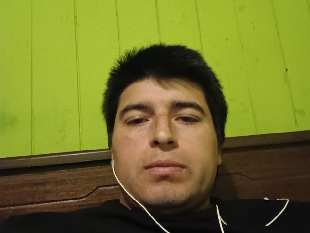 HotChileGuy with short black hair, white earbuds, and a serious face, wearing a black hoodie, against a green wooden wall. His nose is slightly upturned.
