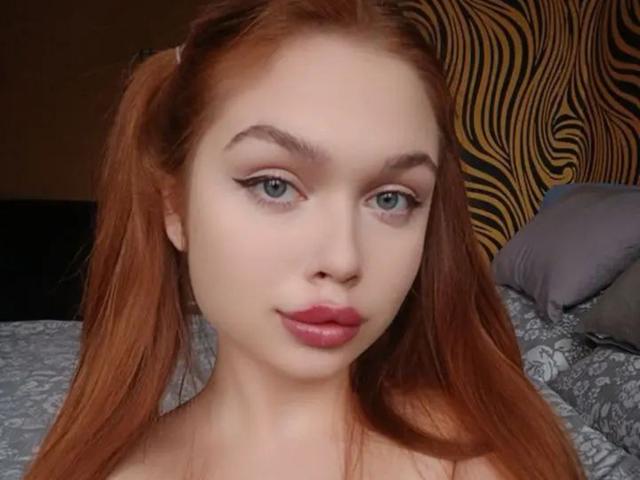 LaraPika has red pigtails, green eyes, and pink lips with subtle makeup. She's staring at the camera with a neutral expression, showing her bare shoulders.