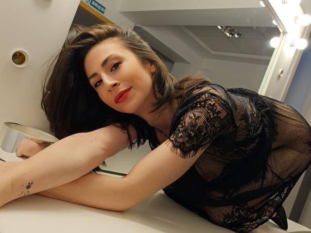 SeductiveAmmy with red lipstick, black lace dress, and visible ass, leaning on a bathroom counter, looking at the camera, brown hair, tattoo on her arm, smiling.