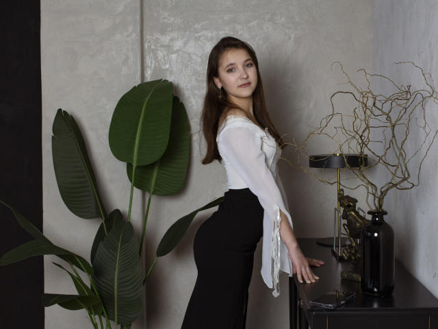 AngelikaFulton with long brown hair, white off-shoulder top, black high-waisted skirt, standing, looking back, hand on dark table, green plant, dry branches in black vase.