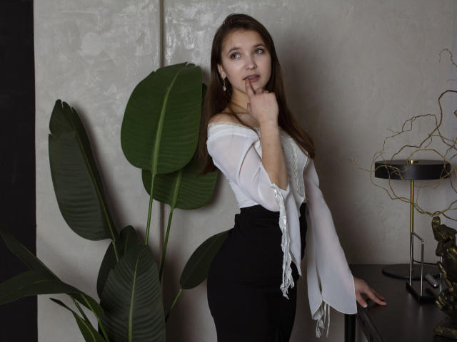 Angelika Fulton, com cabelos castanhos longos, blusa branca com um ombro à mostra, saia preta de cintura alta, dedo na boca, em pé ao lado de uma planta verde, olhando para a câmera, parede cinza ao fundo..