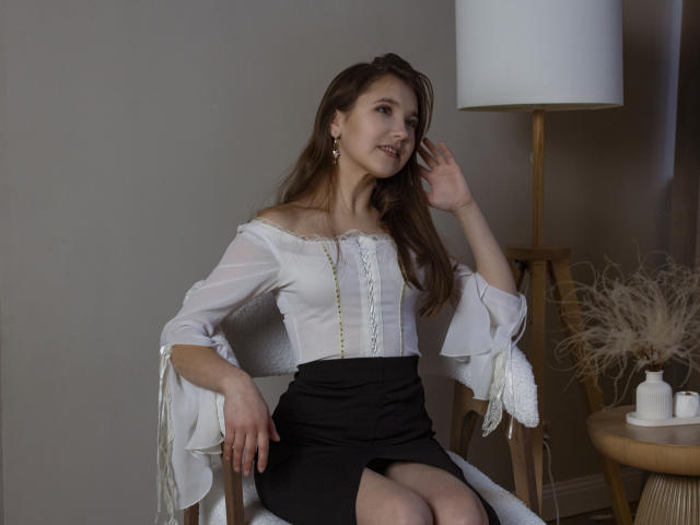 AngelikaFulton with long brown hair, white off-shoulder top, black skirt, and small tits, sitting, hand touching hair, smiling, white lamp, wooden table, and puffy dried grass in background.
