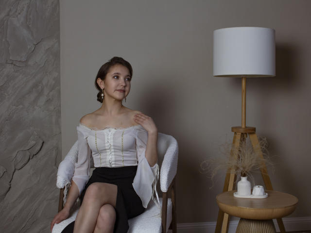 AngelikaFulton with small tits, white off-shoulder blouse, black skirt, sitting on white chair, hand near neck, wooden floor lamp, white lampshade, small table with tea set.