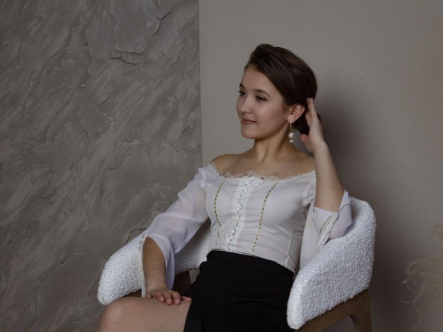 AngelikaFulton with short brown hair, white off-shoulder blouse, black skirt, white textured chair, hand in hair, smiling, earrings, pale skin, cute, small tits, soft lighting.