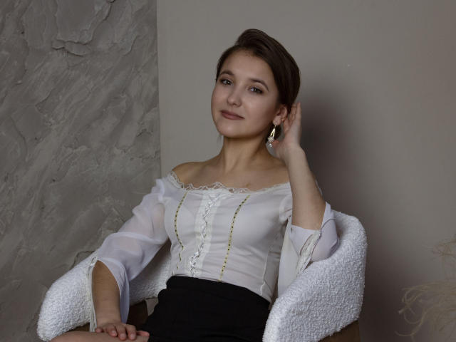 AngelikaFulton with short brown hair, wearing a white off-shoulder top, black skirt, and white textured coat, sits, hand in hair, earrings, soft smile, cute, small tits.