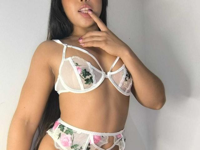 Heyly with her hand on her lips, wearing sheer white floral lingerie, biting her finger while licking her lip.