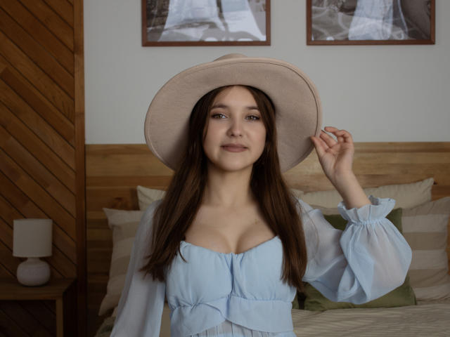 Angelika Fulton, com longos cabelos castanhos, vestindo um vestido azul claro e um chapéu bege, segurando o chapéu com um sorriso suave, exibindo seus seios em uma luz natural e suave..