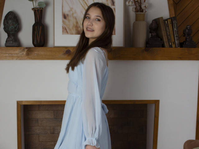 AngelikaFulton, with long brown hair, smiles at the camera, wearing a light blue dress with sheer sleeves, standing in front of a wooden shelf with various decorative items.