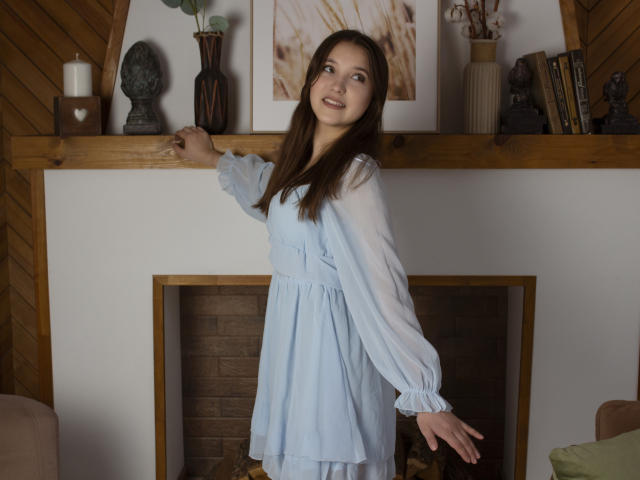 AngelikaFulton stands in a wooden room, wearing a light blue dress, with her right arm on a wooden shelf, brown hair, smiling, tits covered, pussy not visible.