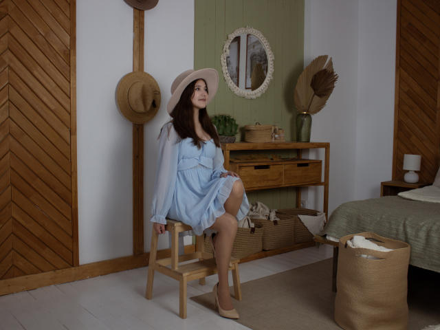 AngelikaFulton with big tits, wearing a light blue dress and beige hat, sitting on a wooden stool, wooden walls, green wall, mirror, baskets, and hat in a rustic room.