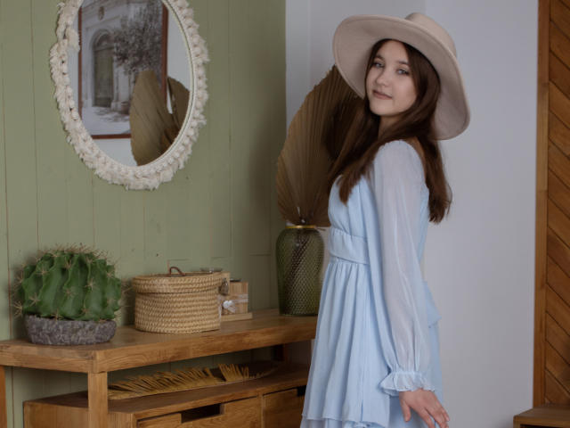 AngelikaFulton with long brown hair, blue dress, and beige hat, standing sideways, smiling, showing her ass, next to a green vase and cactus on a wooden table.