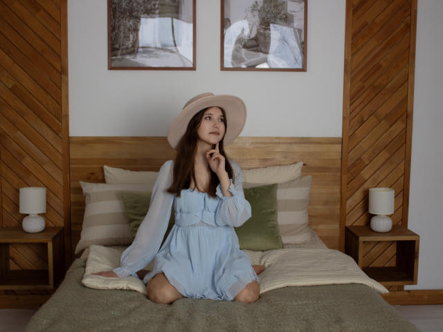 Angelika Fulton, com cabelo castanho, vestindo um vestido azul e um chapéu branco, sentada em uma cama, olhando para cima com uma expressão adorável, seus seios levemente visíveis através do vestido..