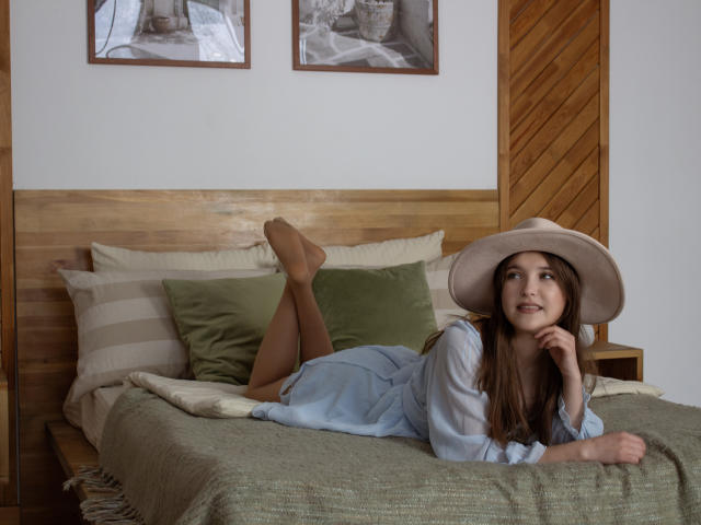 AngelikaFulton laying on a bed, wearing a blue dress, beige hat, and tan stockings, with her legs up, showing her bare ass, looking to the side with a smile.