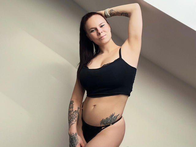 SuesseBertha-hot tiene piel clara, cabello oscuro, tatuajes en los brazos y la parte baja del abdomen, lleva un top negro y una tanga negra, está de pie con un brazo levantado y sonriendo..