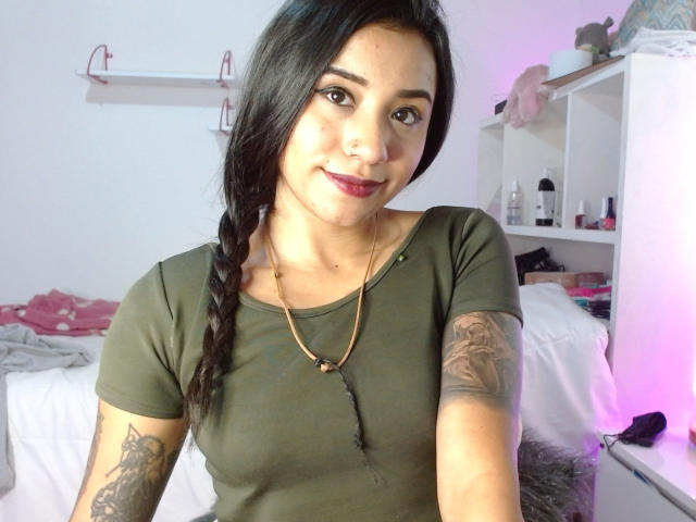 AshleyMayer with dark hair in a braid, green top, tattoos on arms, red lipstick, nose ring, necklace, smiling, standing in a bedroom with white shelves and pink light.
