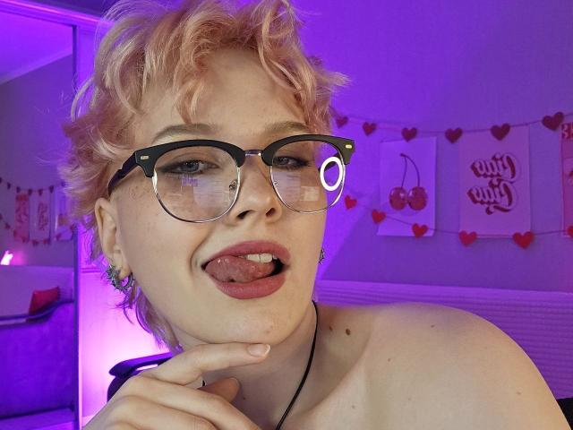 ShellyLove69 with short pink hair, black glasses, and nose ring, tongue out, finger touching chin, no clothes, purple room, heart decorations, and cherry artwork in background.