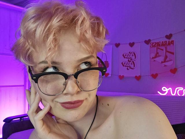 ShellyLove69 with short, curly pink hair, black glasses, freckles, and pink lipstick, touching her cheek, smiling, topless, purple and pink neon lights in the background.