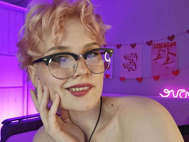 ShellyLove69 with curly pink hair, black-rimmed glasses, and a smile, holding her cheek, showing her bare shoulders and a black necklace. Neon pink light in the background.