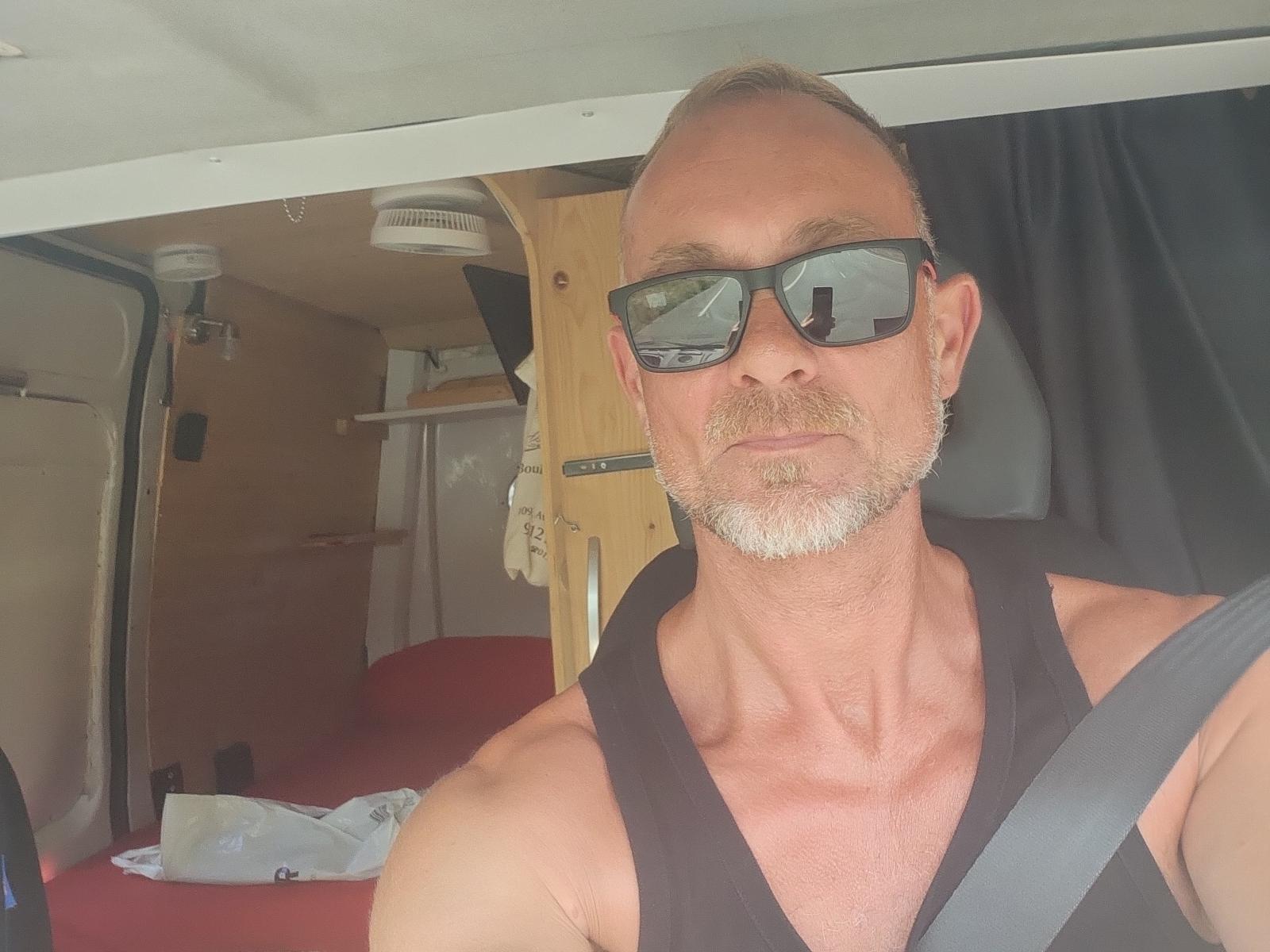 SobenHotX, middle-aged dude with a shaved head, gray beard, black sunglasses, black tank top, seatbelt, and a serious expression, driving in a van.