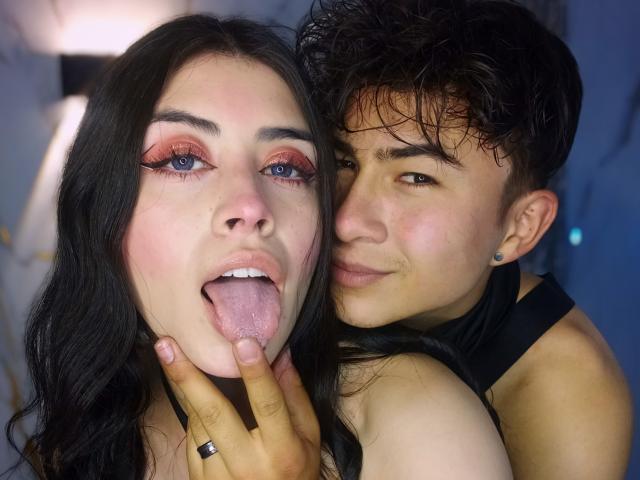 NoahAndRose with long black hair, pink eyeshadow, and tongue out, being gently grabbed by NoahAndRose with short dark hair, black top, and earrings, in a playful pose.