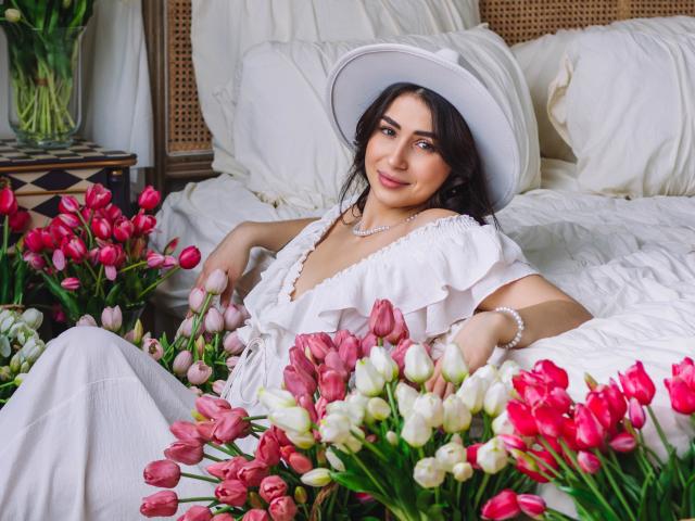 OliviaCluff with dark hair, white dress, and hat, sitting on bed surrounded by pink and white tulips, smiling, pearl necklace, white pillows, and wicker headboard in background.