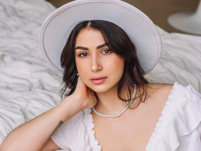 OliviaCluff with dark wavy hair, wearing a white hat and dress, pearl necklace, and looking at the camera, hand on neck, soft skin, and subtle makeup.