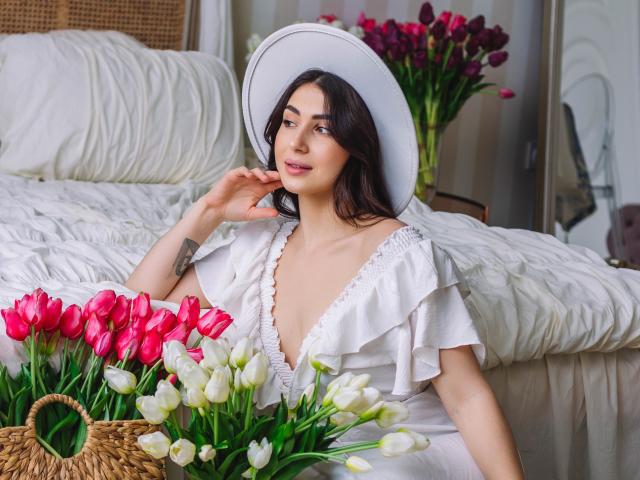 OliviaCluff with dark hair, white ruffled dress, and wide hat, sitting by a bed, holding a bouquet of pink and white tulips, showing a tattoo on her left hand.