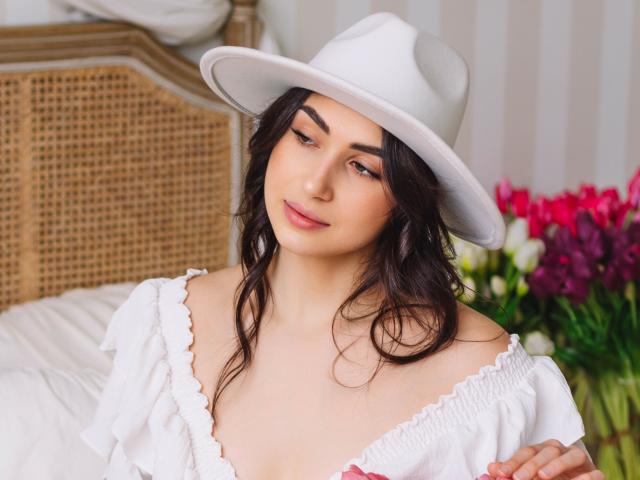OliviaCluff with wavy dark hair, white hat, and off-shoulder white dress, smiling softly, showing her tits slightly, with a wicker headboard and colorful flowers in the background.