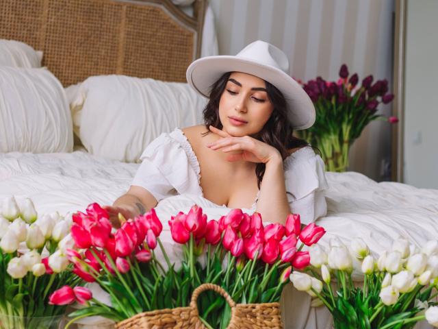 OliviaCluff with white hat, off-shoulder top, and pink-white tulips, resting chin on hand, showing her tits, with a wicker basket in front, in a cozy bedroom.