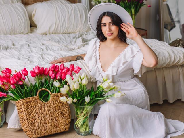 OliviaCluff with medium tits, wearing a white ruffled dress and hat, sitting by a bed with pink and white tulips, wicker basket, and white pillows. Bedroom setting.