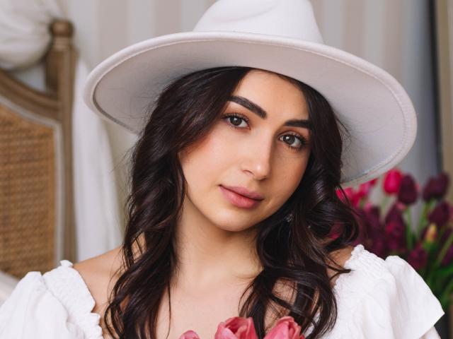 OliviaCluff with dark wavy hair, wearing a white hat and puffy white top, holding pink tulips, has a cute face, brown eyes, and a slight smile.