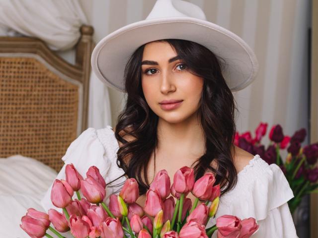OliviaCluff with wavy dark hair, white hat, and off-shoulder white dress, holding pink tulips, has a cute face, and a subtle smile, in a cozy bedroom setting.