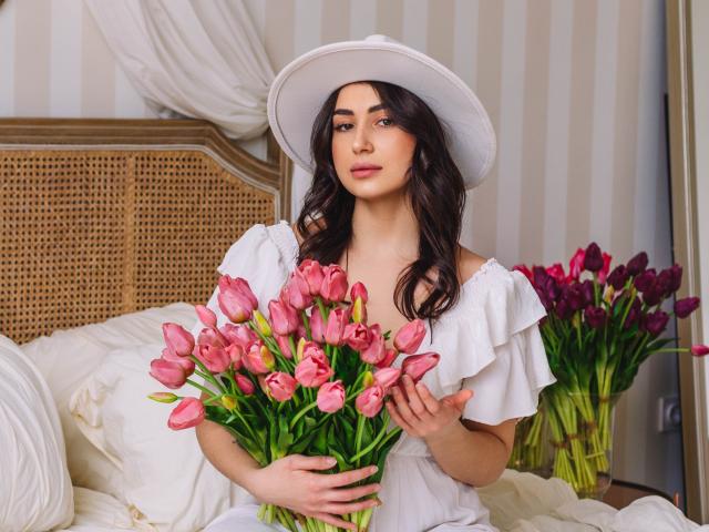 Olivia Cluff, con i capelli castano ondulati, un cappello bianco e un abito con una spalla scoperta, tiene in mano tulipani rosa, seduta su un letto con cuscini bianchi e una testiera in vimini, appare carina e sensuale..
