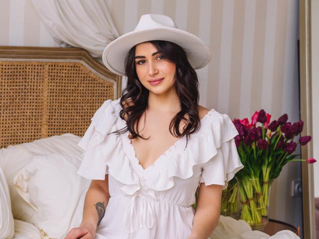 Olivia Cluff, con cabello oscuro y ondulado, vestido blanco con volantes y sombrero blanco, sonriendo, tiene un busto atractivo, y lleva un sombrero de vaquero blanco, está de pie en un dormitorio con tulipanes rosas..