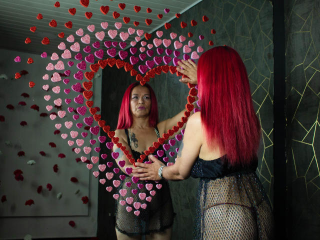 LizhCarthner with red hair, wearing black mesh lingerie, holds a heart-shaped mirror of pink and red hearts, staring at herself, showing her tits and a tattoo on her chest.